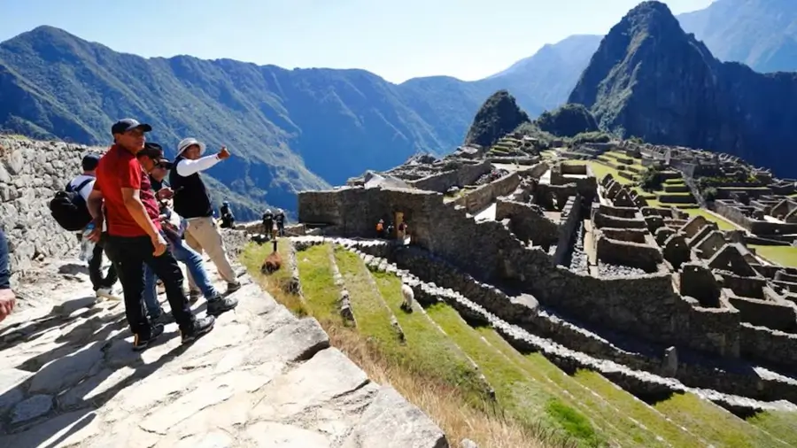 Turistas recorren sendero de piedra y observan las ruinas de Machu Picchu con el Huayna Picchu al fondo.